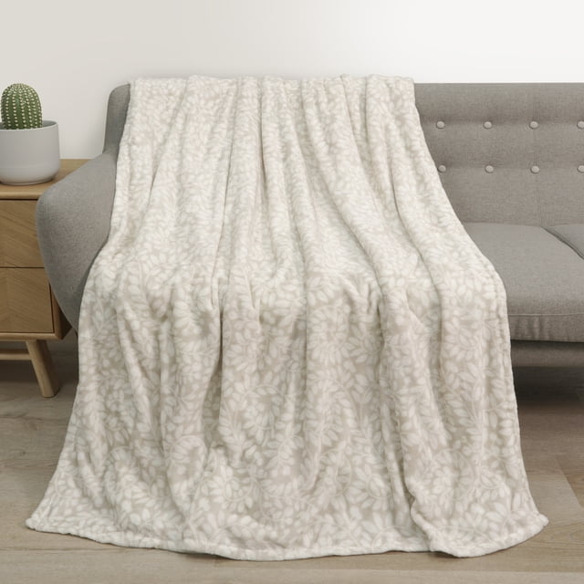 Holiday Time Soft Plush Throw blanket draped over a light grey tufted sofa, featuring a cream base with a white floral pattern. Measuring 50 by 60 inches, this throw is made from 75% polyester and 25% recycled polyester, offering cozy warmth and softness. It is ideal for adding texture and warmth to living spaces.