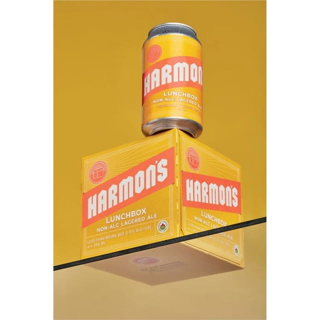 Lunchbox Lager Non Alc 355 ml Canette et Boîte de 4 par Harmon's Craft Brewing, Présentant des Étiquettes Jaunes et orange avec Texte Blanc, Affichées sur une surface Réfléchissante sur Fond Jaune, Mettant en Valeur Cette ale Lager Canadienne Certifiée Biologique Sans Alcool Idéale pour Rafraîchir, Boire Facilement.