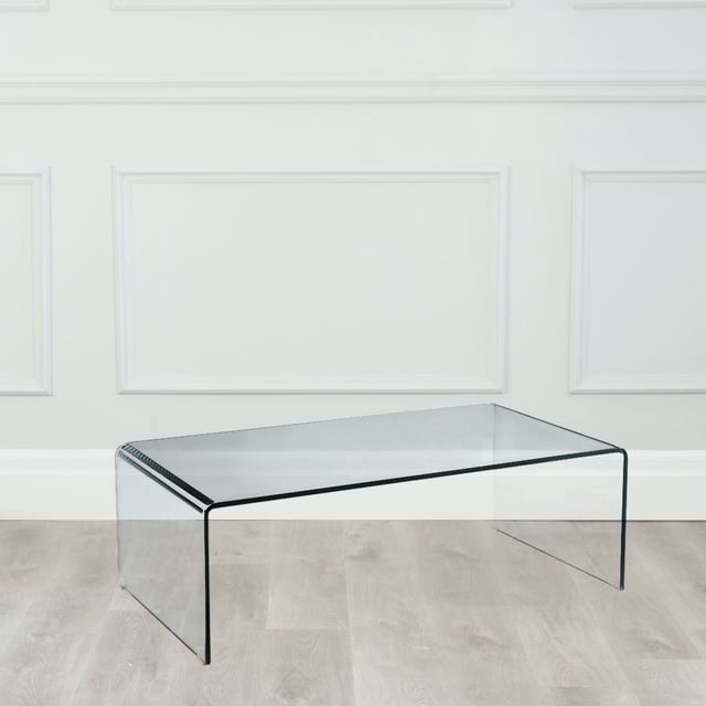 Modern Living Curvo Waterfall Coffee Table crafted from clear tempered glass, featuring a sleek minimalist waterfall design. The durable glass construction is safer than traditional glass and ideal for showcasing rugs or decor in modern homes or apartments. Suitable for small spaces due to its transparent design. Displayed in a bright room with light wood flooring and white panelled walls.