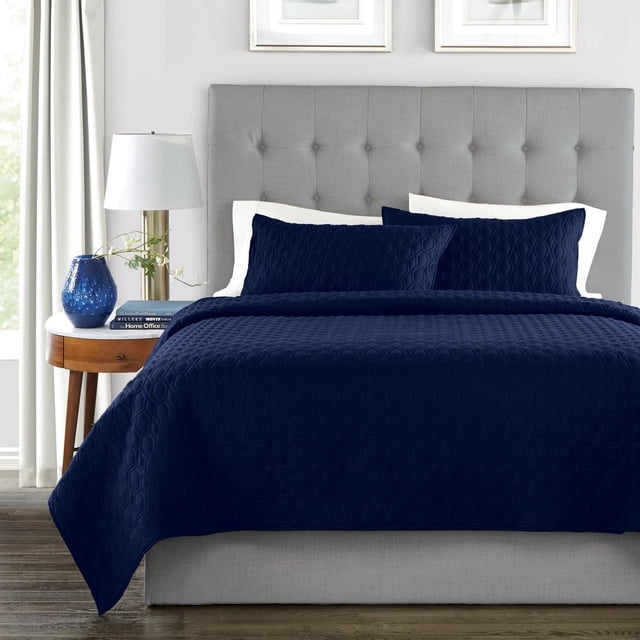 Springmaid Ensemble de Courtepointe Bleu Marine, Couleurs Unies Entretien Facile! Comprenant une Courtepointe Bleu Marine et Deux Taies d'Oreiller Assorties avec Courtepointe Géométrique sur un Lit avec une Tête de Lit Capitonnée Gris Pâle. la Literie Est Superposée sur des Oreillers Blancs dans une Pièce Lumineuse avec une Table de Nuit en Bois Tenant un vase Bleu, une Lampe et des Livres. Cet Ensemble de Courtepointe Polyvalent Est Idéal pour la Maison Ou les Voyages.