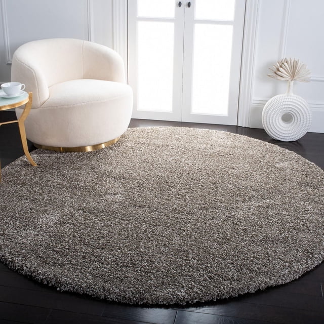 Safavieh Milan Harlow Tapis Shag Uni Gris, un Tapis Shag Rond en Fil Synthétique Pelucheux sur Plancher de Bois Franc Foncé, Montré dans une Pièce Minimaliste Éclatante avec des Murs Blancs, à Côté d'Un Fauteuil Courbé Crème avec une base Dorée et une Petite table d'Appoint Ronde avec une Tasse et une Soucoupe Blanches, Accentué d'Un vase Décoratif Circulaire Blanc avec des Feuilles de Palmier Séchées. Ce Tapis Convient Aussi pour Ajouter de la Chaleur et de la texture aux Espaces Contemporains de la Maison Ou du Bureau.