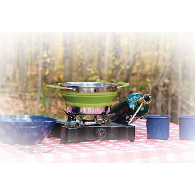 World Famous Collapsible Camping Pot with Lid - Small with green silicone sides and stainless steel top and bottom, sitting on a black portable camping stove on a red and white checkered tablecloth with blue enamel camping mugs and bowl, outdoors in an autumn forest. Its fold-flat design makes it suitable for compact camp kitchen setups.