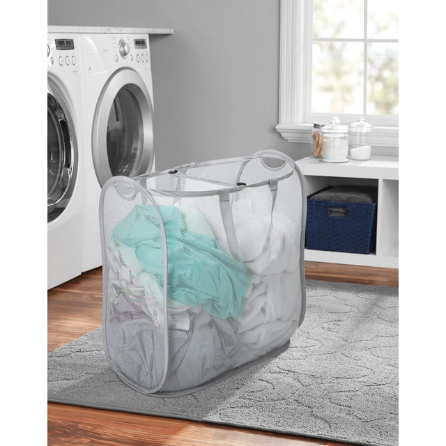 Mainstays Dual Pop-Up Mesh Hamper in grey with breathable mesh fabric and dual compartments filled with laundry, placed on a grey patterned rug in a laundry room with white front-load washer and dryer, white countertop with storage baskets and glass jars, lit by natural window light.