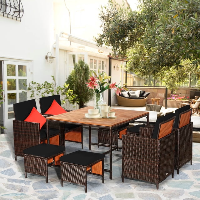 Costway 9 PCS Patio Rattan Dining Set featuring a black PE wicker design with a sturdy steel frame and acacia wood tabletop. The set includes a square table with an umbrella hole, four cushioned armchairs, and four cushioned ottomans, all with removable polyester covers. The chairs and ottomans can be stored compactly under the table. Ideal for outdoor spaces like patios, gardens, or poolside. Shown in a bright outdoor setting with floral decor on the table.