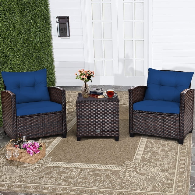 Costway 3PCS Patio Rattan Furniture Set Cushioned Conversation Set Coffee Table Navy. This outdoor patio conversation set includes two rattan armchairs with navy cushions and a square coffee table with a tempered glass top. Perfect for gardens, patios, or poolside, the set is displayed on a patterned beige rug with a white wall and greenery in the background.