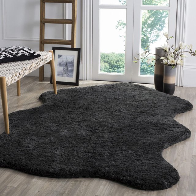 Safavieh Sheep Addie Solid Plush Shag Area Rug in dark charcoal gray with a 1-inch plush shag pile and organic irregular shape, placed on light wood flooring near a woven bench with a black and white geometric pillow, a wooden ladder, framed black and white photo, and a tall vase with white flowers by a glass door, often used to add warmth and texture to accent rug areas.