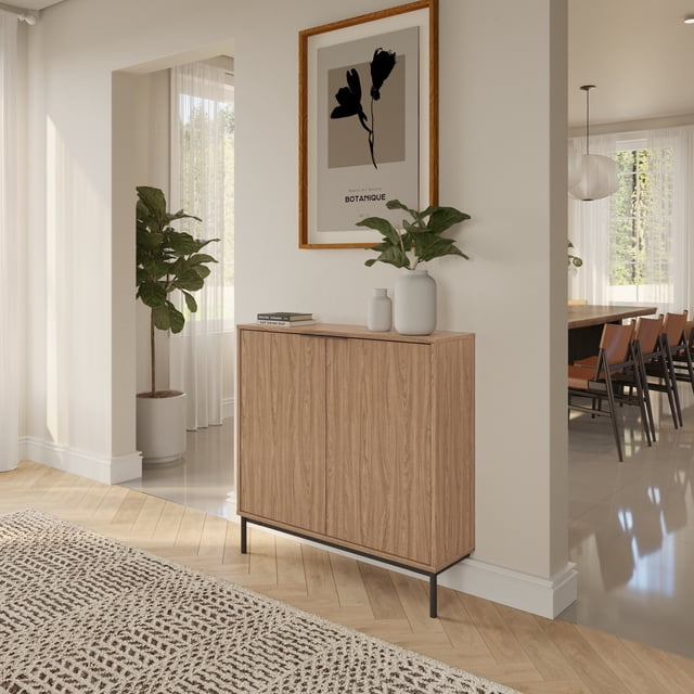 Nexera Slim 2-Door Storage Console in light walnut with black metal hairpin legs, featuring a slim, wall-mounted design ideal for narrow spaces, two soft-close doors concealing internal shelves, styled with white vases and a green plant in a bright room with wood parquet flooring.