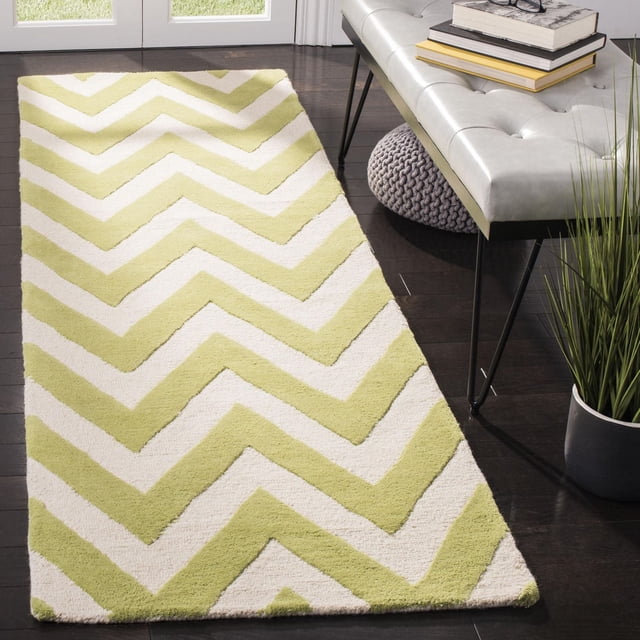Safavieh Cambridge Kaitlyn Zig Zag Stripes Area Rug in hand-tufted wool with a light green and cream zig zag pattern, placed on dark hardwood flooring beside a grey tufted bench and a knitted pouf, suitable for enhancing modern home or office interiors with durable texture and style.