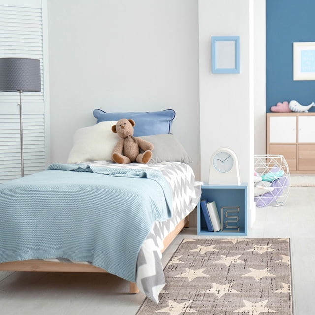ECARPET Kids Room, Nursery, Playroom, Bedroom Area Rug, Angel Collection with a grey star-patterned rug placed beside a wooden bed with blue and grey bedding and a teddy bear, accompanied by a blue nightstand with a clock and books, crafted for softness and durability in high-traffic kids' rooms, also suitable for nursery or playroom decor.