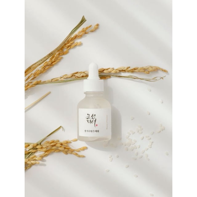Beauty of Joseon Glow Deep Serum Riz + Arbutine 30 ml dans une Bouteille Compte-Gouttes en Verre Givré avec Bouchon Blanc, Sertie sur Fond Blanc avec des Tiges de Riz Séchées et des grains de Riz Éparpillés, Mettant en Valeur Sa Formule à Base d'Eau Qui Illumine, Unifie le Teint et Hydrate la Peau Sèche.