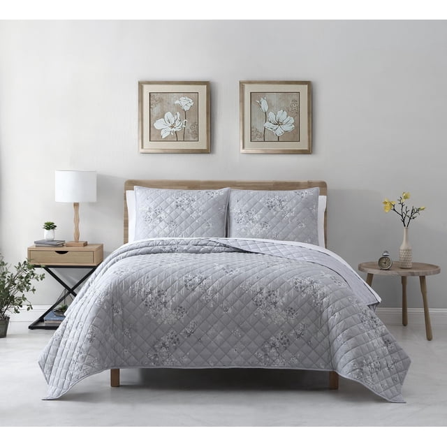 Avery Homegrown Ensemble de Courtepointe Grise à Motif Floral 3 Pcs sur un Lit en Bois avec une Courtepointe Grise et Deux Taies d'Oreiller Assorties Présentant de Subtils Motifs Floraux et une Courtepointe à Losanges. Fait de lyocell et de Coton, Absorbe l'Humidité, Régule la Température et Reste Frais. la Chambre Comprend une table de Nuit en Bois avec une Lampe Blanche et une Plante, une table d'Appoint Ronde avec un vase de Fleurs Jaunes, et Deux Imprimés Floraux Encadrés au-Dessus du Lit.