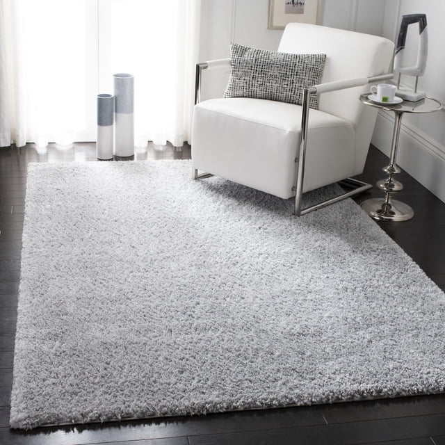 SAFAVIEH August Carlene Solid Plush Shag Area Rug in light grey on dark hardwood floors, paired with a white leather armchair with metal frame and patterned lumbar pillow, next to a round silver side table holding a white cup and modern sculpture, showcasing the rug's plush polypropylene yarn texture and casual West Coast aesthetic, often used to add warmth and softness to living rooms.