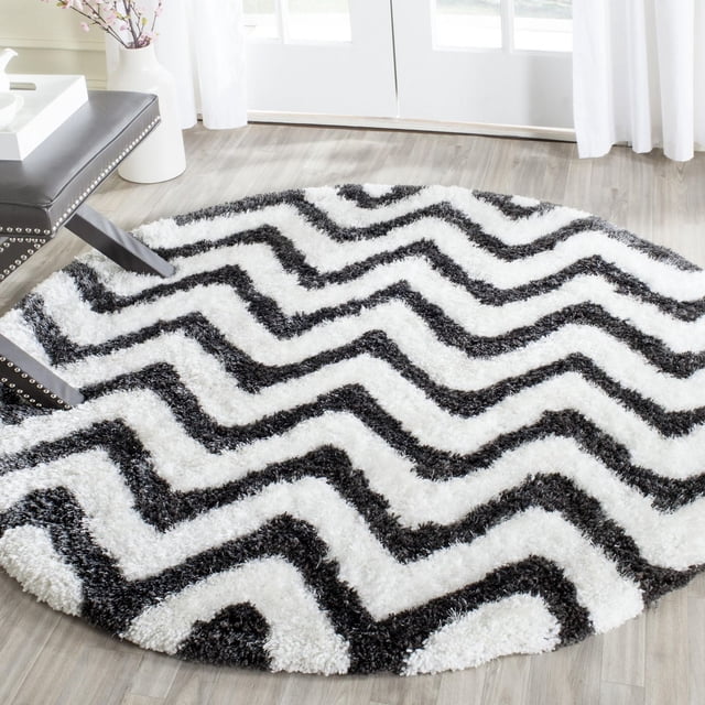 Safavieh Barcelona Carmella Zigzag Stripes Shag Area Rug, round with black and white zigzag stripes and plush shag texture, displayed on light wood flooring near a black studded bench and white vase with flowers by glass doors with white curtains, ideal for modern accent rugs in living rooms.