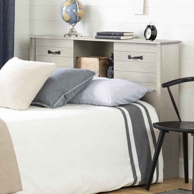 South Shore Ulysses Full Bookcase Headboard with Doors in winter oak finish, featuring two sliding doors with black handles and three open storage compartments for bedside storage. Shown with a bed dressed in white and grey striped bedding and pillows, the headboard holds a globe, books, and an alarm clock on top. Positioned beside a black chair against a white shiplap wall, it offers practical bedroom furniture storage and cable management.