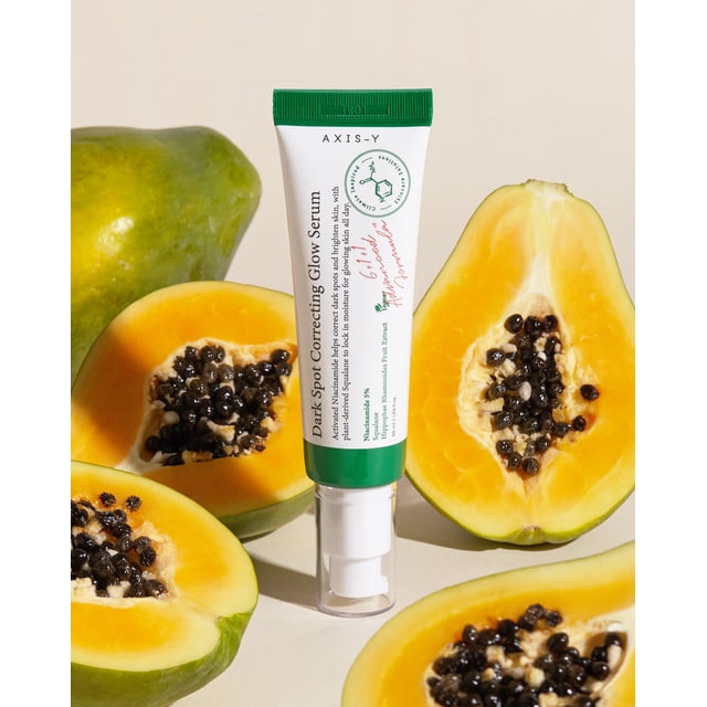 AXIS-Y Dark Spot Correcting Glow Serum, a vegan facial serum in a white and green tube, displayed upright among halved papayas with visible seeds. Formulated with 5% niacinamide to reduce dark spots and refine skin texture, and rice extract for hydration. Artificial fragrance-free.