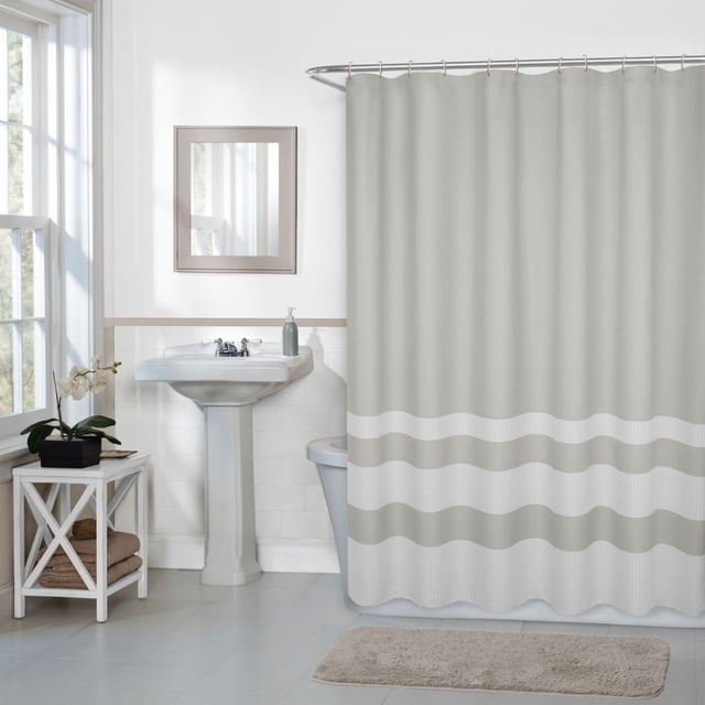 Hometrends Textured Waffle Stripe Fabric Shower Curtain in light beige with off-white two-toned waffle stripes, hanging from a curved metal rod with buttonhole top. Shown in a bathroom with a white pedestal sink, a small white side table with folded towels and a potted orchid, and a beige bath mat on tiled floor. Suitable for use with a liner to protect against water.