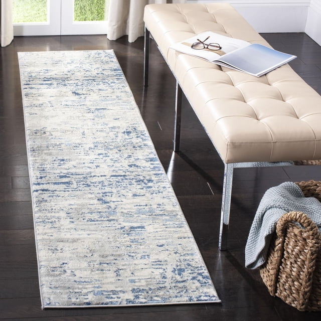 SAFAVIEH Jasper Brandt Abstract Overdyed Area Rug runner with soft cut pile in a faded abstract pattern of blue and cream tones, placed on dark hardwood flooring beside a beige tufted bench with chrome legs and a woven basket with a light blue throw, ideal for contemporary narrow spaces.