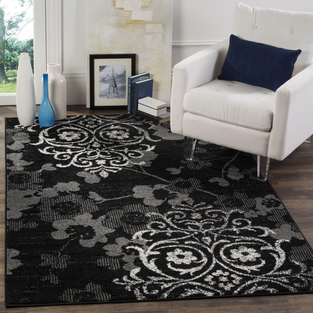 SAFAVIEH Adirondack Hortense Floral Area Rug in black and grey with detailed floral and scroll designs, made of durable polypropylene yarns, placed on dark hardwood floor beside a white armchair with a navy blue pillow, accented by decorative vases and framed artwork, ideal for enhancing living room decor.