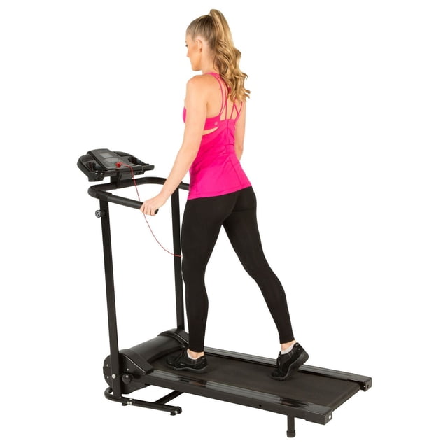 Fitness Reality TRE2500 Folding Electric Treadmill with Goal Setting Computer showing a woman walking on the treadmill wearing a pink tank top and black leggings, holding black handlebars with a safety clip attached, featuring a backlit LCD display for time, distance, calories, and speed, and a compact folding design suitable for small spaces.