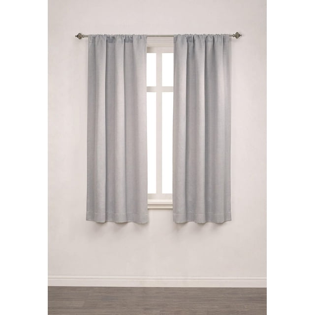 Mainstays Chambray Blackout 63" Window Curtains Panel Pair in soft silver heather texture, hanging on a metal rod over a white-trimmed window in a neutral room with wood flooring. These 100% polyester blackout curtains feature triple foam backing and rod pocket design for full light blocking, ideal for bedrooms or media rooms.