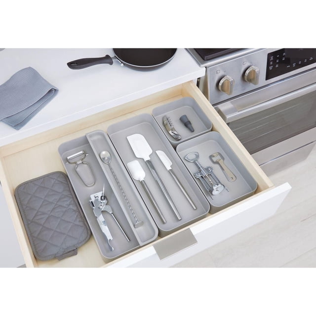 iDesign Gray Eco BPA-Free Recycled Plastic 6" Kitchen Drawer Organizer Bin inside a light wood kitchen drawer, neatly organizing kitchen utensils including spatulas, corkscrew, bottle opener, peeler, and mixing spoon in multiple gray bins, with a gray quilted pot holder, adjacent to a stainless steel stove and white countertop.