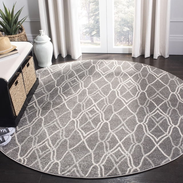 Safavieh Amherst Virginia Geometric Area Rug, round with grey polypropylene cut pile and white geometric lattice pattern, placed on dark hardwood floor by a window with beige curtains, styled with a black bench with woven baskets and a green plant, ideal for durable indoor-outdoor use in high-traffic spaces.