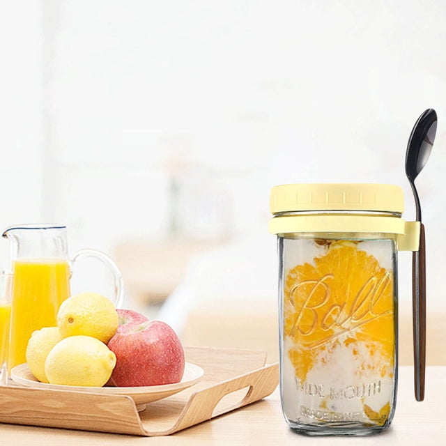 Kcavykas Kitchen overnight oats container with lid and spoon, featuring a 350ML capacity. The portable mason jar is shown with a yellow lid and a black spoon attached to the side. The jar contains layered oats and orange slices, placed on a kitchen counter next to a tray with lemons, apples, and a glass of orange juice.