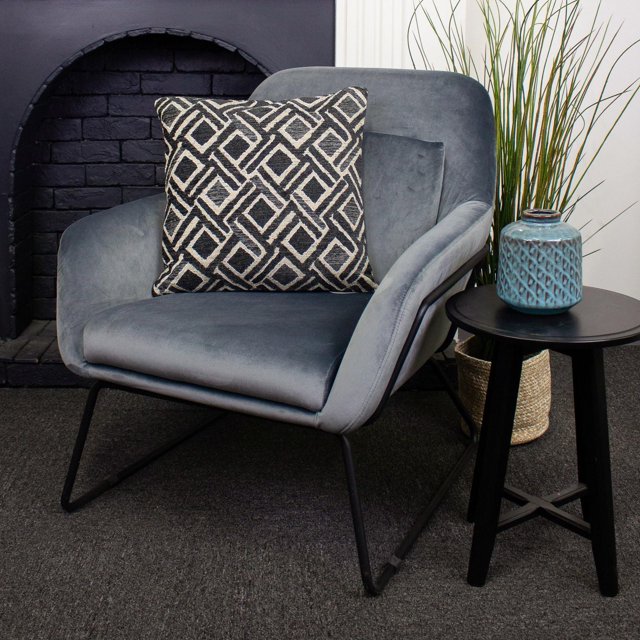 Coussin de Luxe Sabar avec Motif Géométrique Noir et beige sur Fauteuil en Velours Gris avec Pattes en Métal Noires, à Côté d'Une table d'Appoint Ronde Noire Tenant un vase en Céramique Texturé Bleu et un Panier Tressé avec une Haute Herbe Verte, devant un Foyer en Briques Noires.