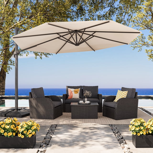 CorLiving 11.5ft Deluxe Tilting and Rotating Offset Patio Umbrella with aluminum frame and warm white canopy, shading a dark grey woven outdoor seating set with two armchairs, loveseat, and coffee table on a beige rug by a pool with ocean view and yellow flowers in black planters.