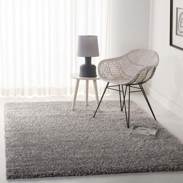 Safavieh California Tapis Shag en Peluche Uni Gris Pâle avec une pile de Polypropylène dense de 4,5 Kg/m2, Exposé dans une Pièce Minimaliste avec une Chaise en Rotin Tressé sur des Pattes en Métal Noires, une Petite table d'Appoint Ronde Blanche avec une Lampe Grise, et un magazine sur le Tapis Près de la Chaise, contre des Rideaux et des Murs Blancs. la texture Peluche Rug's Convient pour Ajouter Confort et Chaleur aux Espaces de Vie.