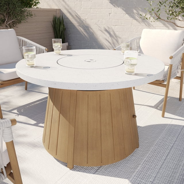 Better Homes & Gardens Steel Propane Fire Pit Table with Glass Rocks & Protective Cover, Brown, shown on a sunlit outdoor patio with a round white speckled tabletop and brown wood-textured steel base. Four glasses with drinks sit on the table, surrounded by cushioned white rope chairs. The fire pit has a central lid that converts the 55,000 BTU propane burner into a table, offering adjustable outdoor heating and ambiance for patios or decks.