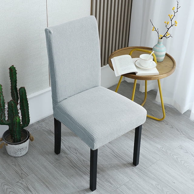 Clearance! Seat Cover Dining Chair Chair Cover Spring Chair Covers Birthday Chair Cover Chairs Covers for Dining Table Seat Chair Covers Slipcovers for Chairs. A dining chair with a fitted slipcover in a light grey textured fabric, featuring an elastic band design for a secure fit. The chair is placed in a modern room with a wooden floor, a small round side table holding a cup and an open book, and a decorative vase with yellow flowers. A potted cactus is visible in the background.