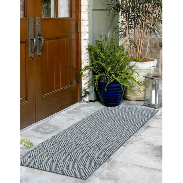 ECARPET Laura Contemporary Runner Rug with a geometric pattern of overlapping angled shapes in azure blue and off-white, made from stain-resistant polypropylene fibers. It is placed on a stone floor by wooden double doors, styled with potted ferns and a metal candle lantern, suitable for indoor and outdoor use.