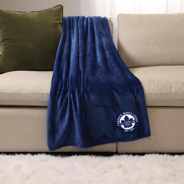 NHL Blue Sports Throw Blanket, 50" x 60", plush deep blue 100% polyester blanket with Toronto Maple Leafs logo embroidered near the bottom edge, draped over a beige sofa with a green cushion, ideal for indoor and outdoor warmth and comfort.