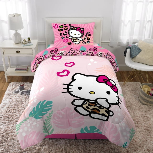Hello Kitty "Wild Jungle" Twin/Full Comforter & Pillowcase set featuring a reversible microfiber comforter with a large Hello Kitty character wearing a leopard print outfit and pink bow on a pink background with tropical leaves and heart accents, displayed on a twin bed in a bright kids' bedroom with white curtains and a nightstand with a lamp.