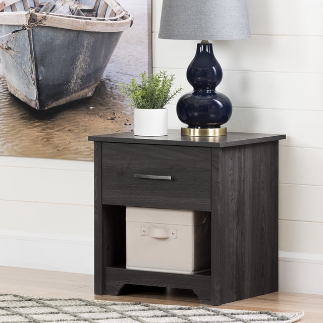 South Shore Fusion 1-Drawer Nightstand in gray oak with a metal handle drawer and open storage holding a beige fabric box, topped with a navy blue lamp with gray shade and a small green plant, set against a white shiplap wall and a large photo of a rustic boat on sand, suitable for bedside storage and organization.