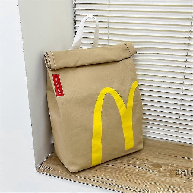 Realistic Mcdonalds Backpack/messenger Side Bag featuring a beige canvas material with a large yellow McDonald's logo on the front. The bag has a roll-top closure, a white handle, and a red side tag. Designed with a large-capacity interior and adjustable shoulder strap, it is suitable for travel or daily use. Positioned on a wooden surface near a window with blinds.