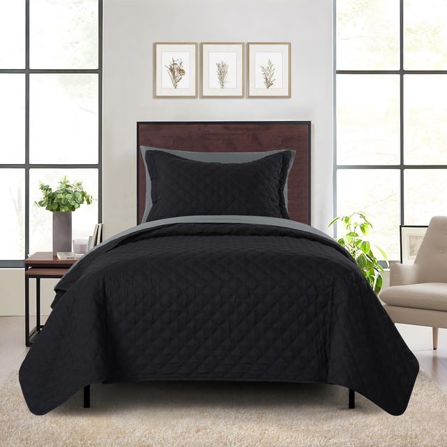 Mainstays quilt-in-a-bag bedding set in black on a twin bed with a dark wood headboard, featuring a quilted black quilt, matching pillow sham, and grey fitted sheet, in a bright room with large windows, beige armchair, side table with books and a plant, and three framed botanical prints above the bed.