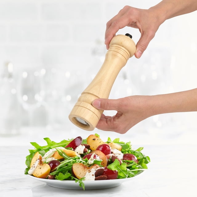 Trudeau 8" Capri Natural Wood Pepper Mill & Salt Mill set with corrosion-resistant ceramic grinder being used to season a fresh salad of leafy greens, sliced peaches, grapes, and creamy dressing on a white plate in a bright kitchen, suitable for adjustable fine to coarse grinding.