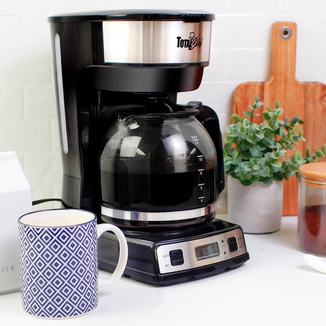 Total Chef 12-Cup Programmable Drip Coffee Maker with Glass Carafe and LCD Display on a white kitchen countertop, featuring a black and stainless steel body, a glass carafe filled with brewed coffee marked up to 12 cups, digital timer on the base, and a blue and white patterned ceramic mug beside it with kitchen decor in the background.