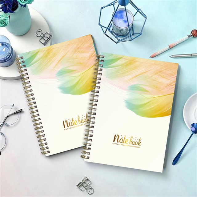 TopLLC English Schedule A5 Plan Book with Separating Pages Plan Notebook Coil Book featuring a navy blue floral pattern with gold double-wire binding. The notebook includes monthly tabs, interior pockets, and ample weekly and monthly pages for planning. Displayed are two notebooks with a colourful feather design on the cover, placed on a light blue desk alongside office supplies like glasses, a pen, and decorative items.
