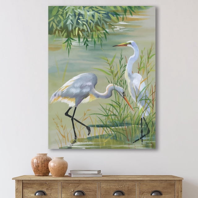 Designart Heron Birds I Canvas Wall Art depicting two white heron birds in green marsh grasses by water, printed on gallery-wrapped premium cotton canvas in traditional style, suitable for home wall décor, displayed above a wooden console table with vases and books.
