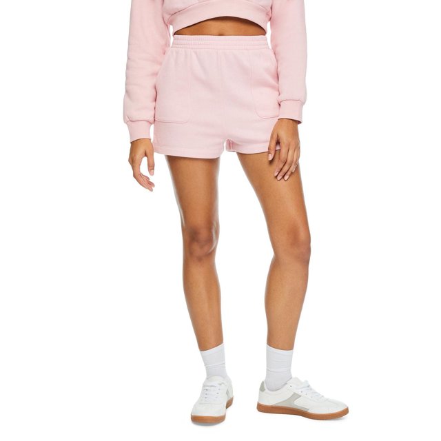 No Boundaries Women's Fleece Short in light pink shown front-facing on a model wearing white socks and white sneakers, featuring an elasticized waistband, two front patch pockets, and split hems for ease of movement, made from soft fleece knit fabric ideal for casual wear or lounging.