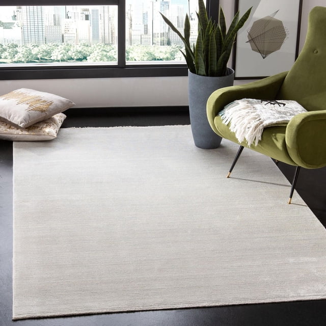 SAFAVIEH Dream Ethelyn Abstract Overdyed Area Rug in a modern room, featuring a large light beige textured rug with subtle linear patterns on a dark floor, complemented by a green upholstered armchair with a fringed throw and glasses, a tall gray planter with snake plants, and decorative pillows near a window overlooking a cityscape, ideal for adding elegant accent and warmth to home décor.