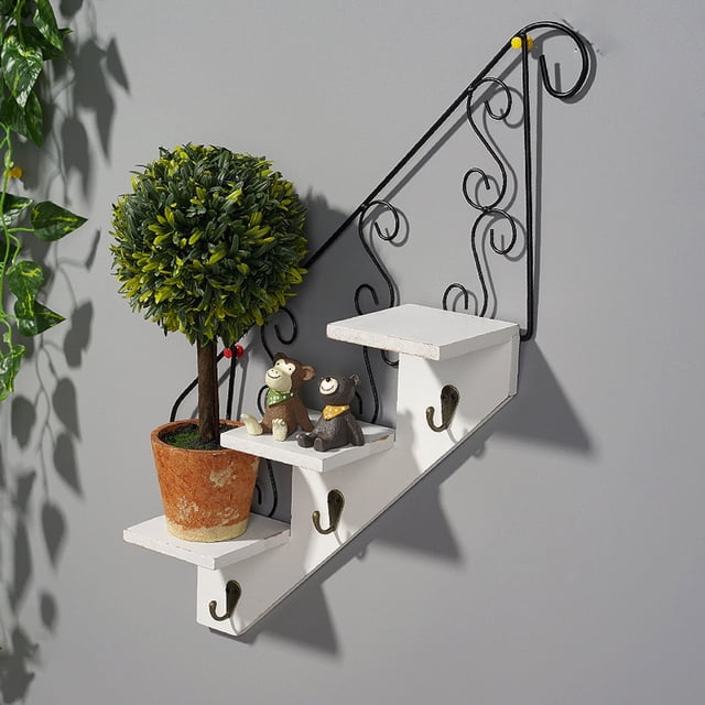 TIMIFIS Iron Rack, a wall-mounted floating shelf with a step-like design, featuring three white wooden tiers supported by black iron scrollwork. Each tier includes a hook underneath for hanging items like keys or hats. The shelf is styled with a small potted plant and decorative figurines, suitable for living rooms, bedrooms, or offices. Dimensions are 45x24cm (17.72x9.45in).