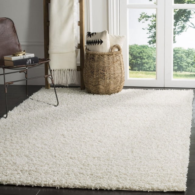 Safavieh Athens Solid Plush Shag Area Rug in soft white with a dense Euro-style shag pile, placed on dark hardwood floors in a bright room. The rug adds plush comfort and warmth, shown with a woven basket holding pillows and a metal chair with books, suitable for enhancing living room decor.