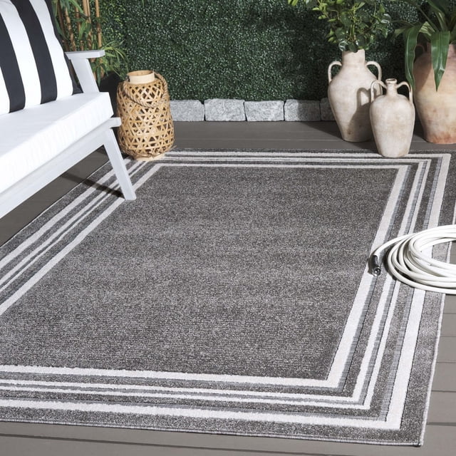 SAFAVIEH Cabana Rado Bordered Indoor/Outdoor Area Rug with grey textured center and white geometric borders, placed on a wooden deck beside a white cushioned chair, woven lantern, coiled white garden hose, and large ceramic vases against a green hedge, suitable for durable indoor or outdoor use.