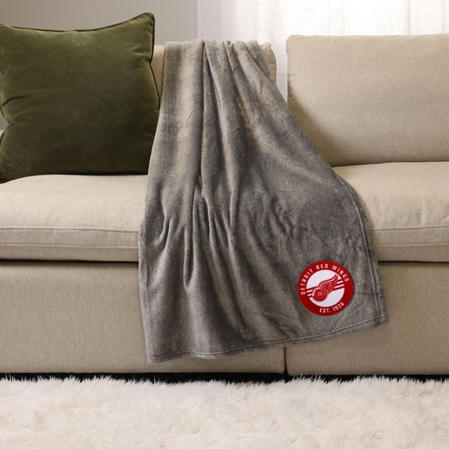 NHL, NBA, MLB Grey Sports Throw Blanket, 50" x 60", draped over a beige sofa with a green cushion, featuring a red Detroit Red Wings logo patch, made of soft plush polyester for warmth and comfort, suitable for indoor and outdoor use.