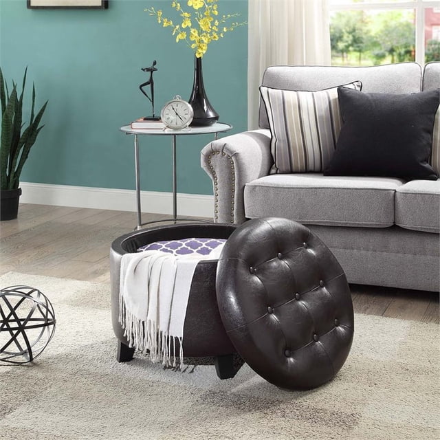 Convenience Concepts Designs4Comfort Rond Ottoman en Similicuir Espresso Exposé dans un Décor de Salon. le Ottoman Présente un Couvercle Capitonné Amovible, un Rangement Dissimulé à l'Intérieur, Idéal pour Ranger des Couvertures Ou des magazines. Rembourré en Similicuir Espresso avec des Pattes en Bois Massif, Il Complète la Déco Environnante, Incluant un sofa Gris et une table d'Appoint. Convient comme Ottoman de Rangement Ou Pièce Décorative d'Accent.