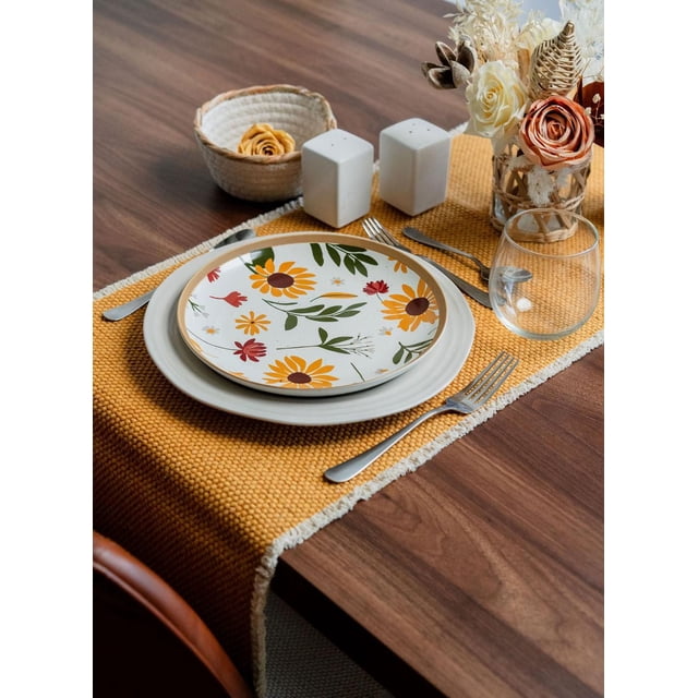 Fabstyles 100% Cotton Bali Table Runner in mustard yellow with white fringe edges on a dark wood dining table, featuring a place setting with a white dinner plate topped by a floral plate with yellow and red flowers, stainless steel cutlery, clear stemless glass, white salt and pepper shakers, and a vase of dried and fresh flowers including roses and pine cones, suitable for indoor and outdoor use.